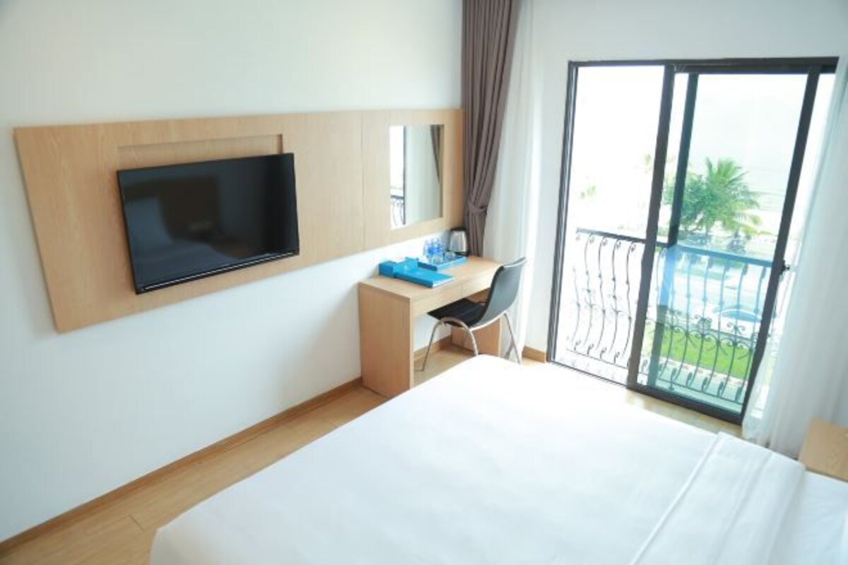 A hotel room with a bed, a desk with a chair, a television mounted on the wall, and a balcony with a view of palm trees.