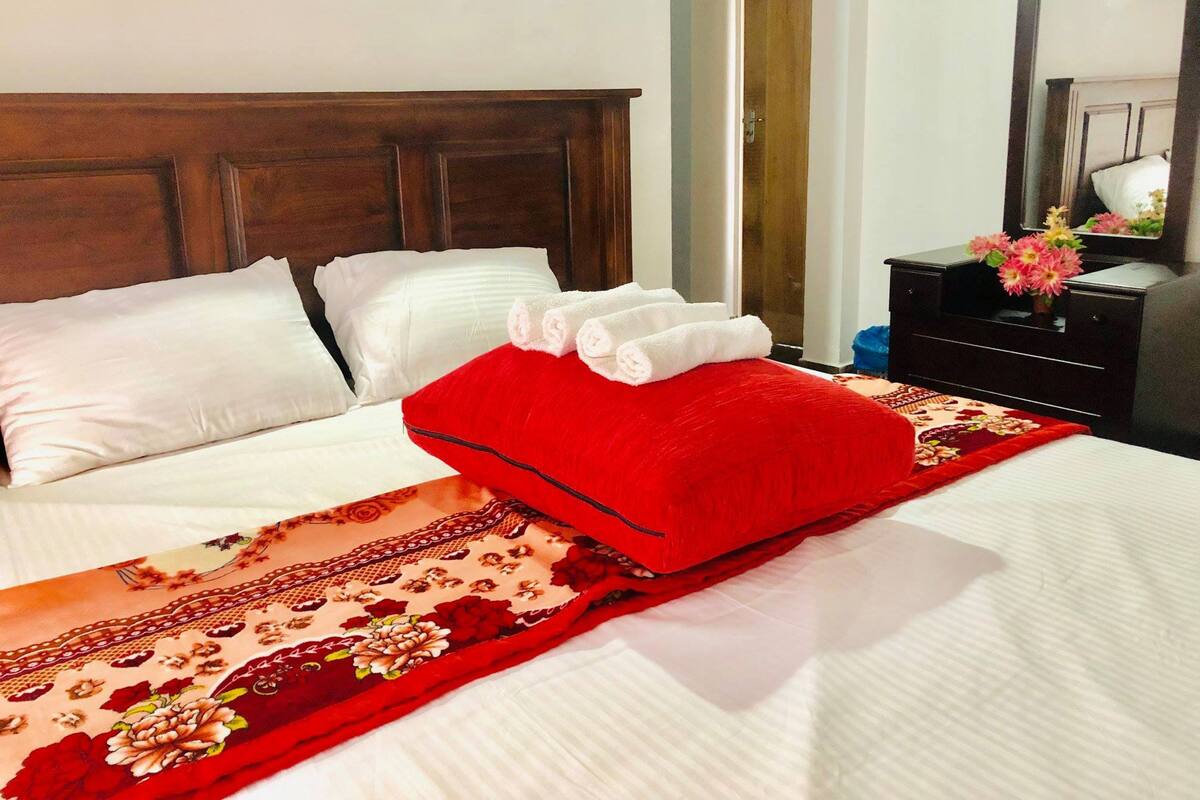 A neatly made bed with white linens, a red pillow, and a floral-patterned bedspread.