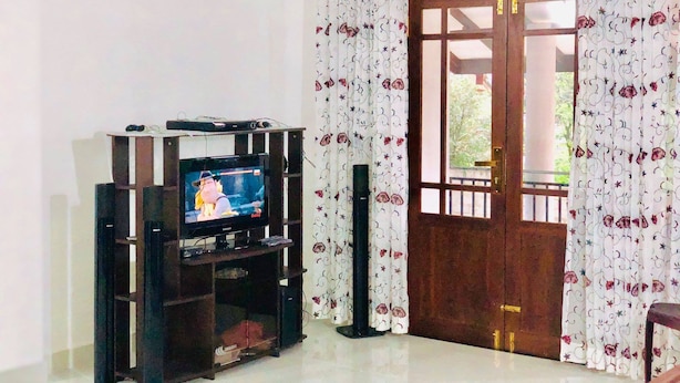 A living room with a TV on a wooden stand, a door with a wooden lattice design, and floral curtains. - Family Ties GUEST INN (Bandarawela)