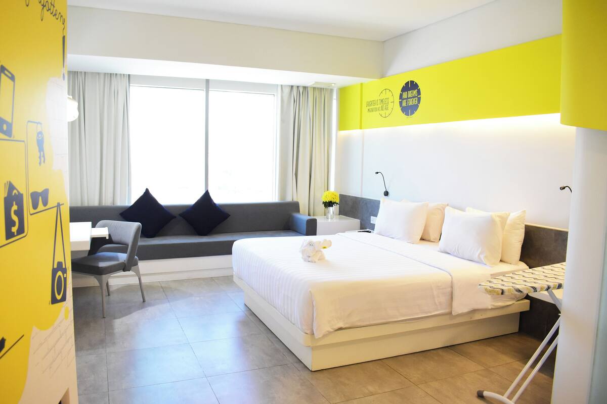 A hotel room with a large bed, a sofa, and a chair. There is a window with curtains, a wall with a yellow sign, and a lamp on a bedside table.