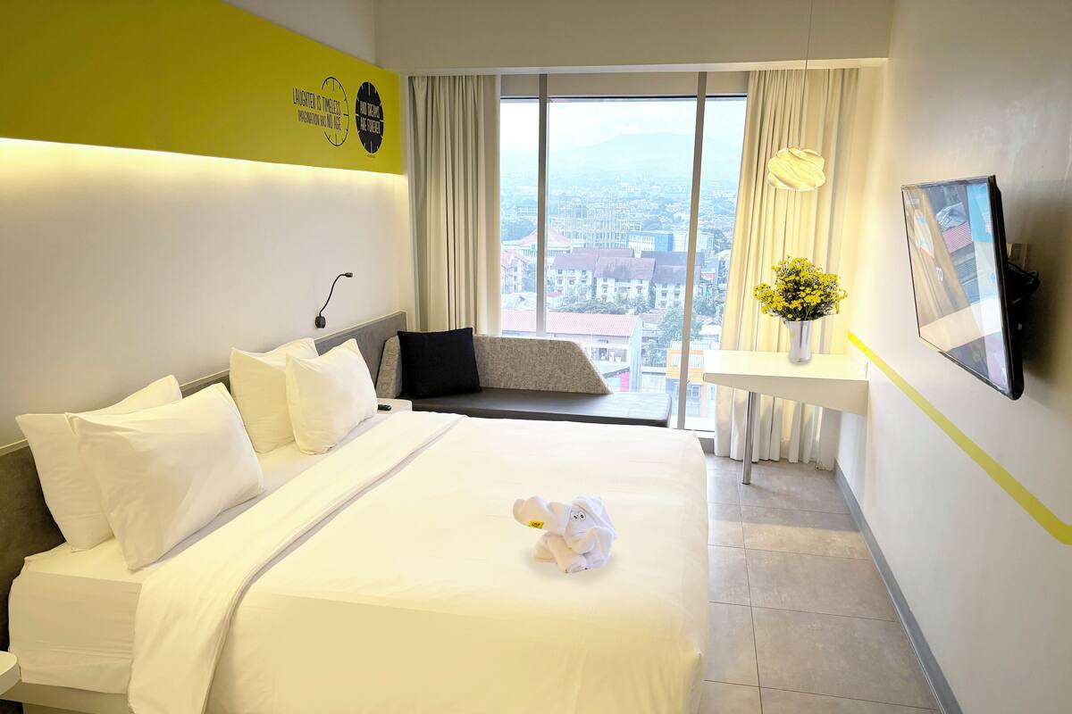 A hotel room with a large bed, a desk, a TV, and a view of the city through the window.