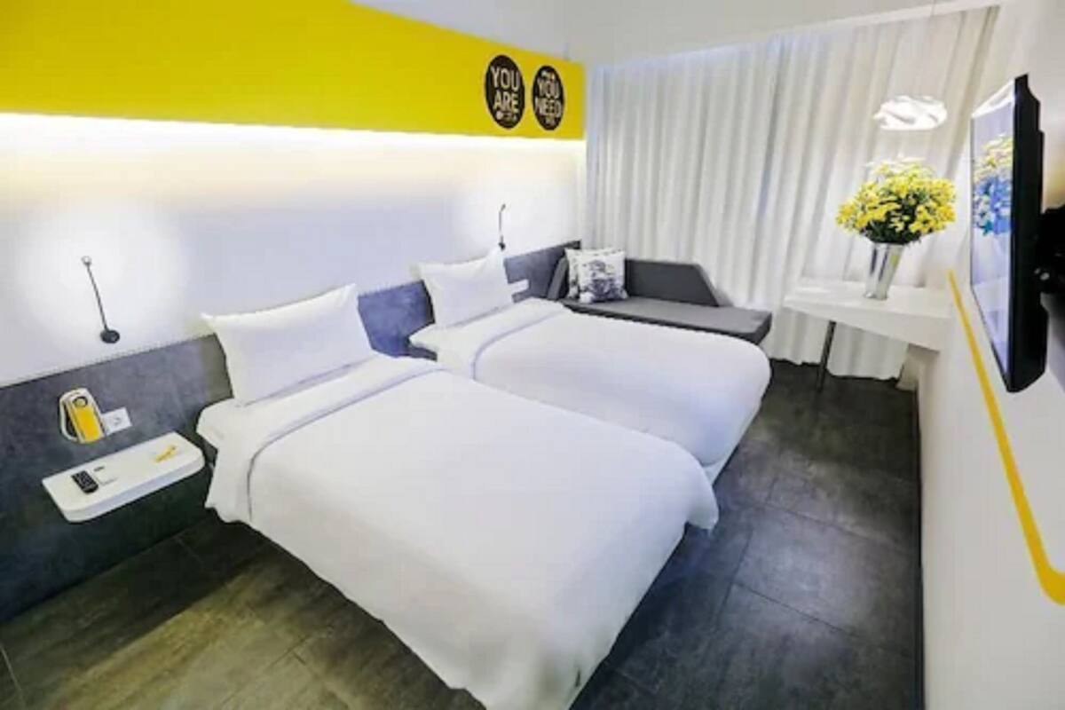 A hotel room with two beds, a small desk, a TV, and a wall-mounted light fixture.