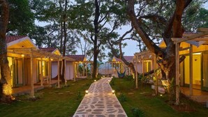 A pathway leading to a row of wooden cabins with glass doors, surrounded by trees and a well-maintained lawn. - Blu Resorts (Vagator)