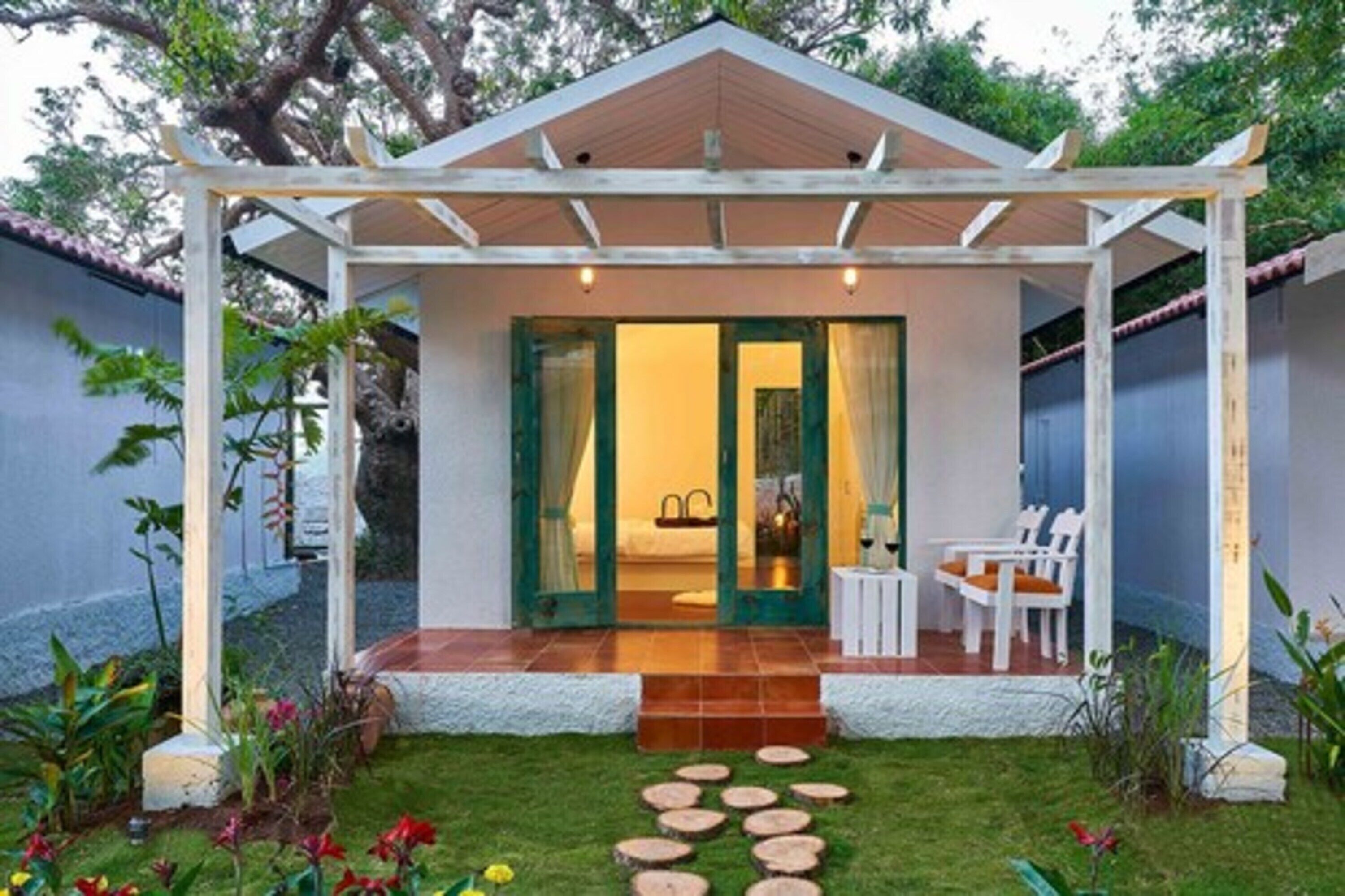 A covered outdoor seating area with a patio, a small garden with stepping stones, and a view into a room with a bed and a sink.