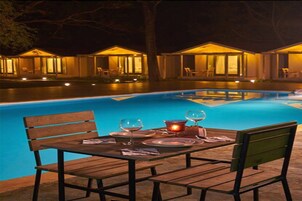 Zona de comedor junto a la piscina con una mesa dispuesta para dos, rodeada por una piscina y un edificio con varias ventanas.