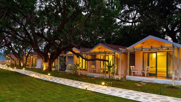 A row of wooden cabins with large windows, surrounded by trees and a stone pathway. - Blu Resorts (Vagator)