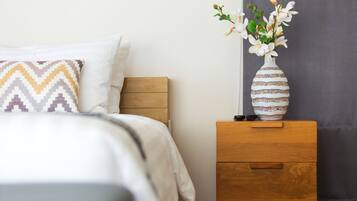 Una cama bien hecha con un cojín estampado, una mesita de noche con un florero y una cortina oscura.