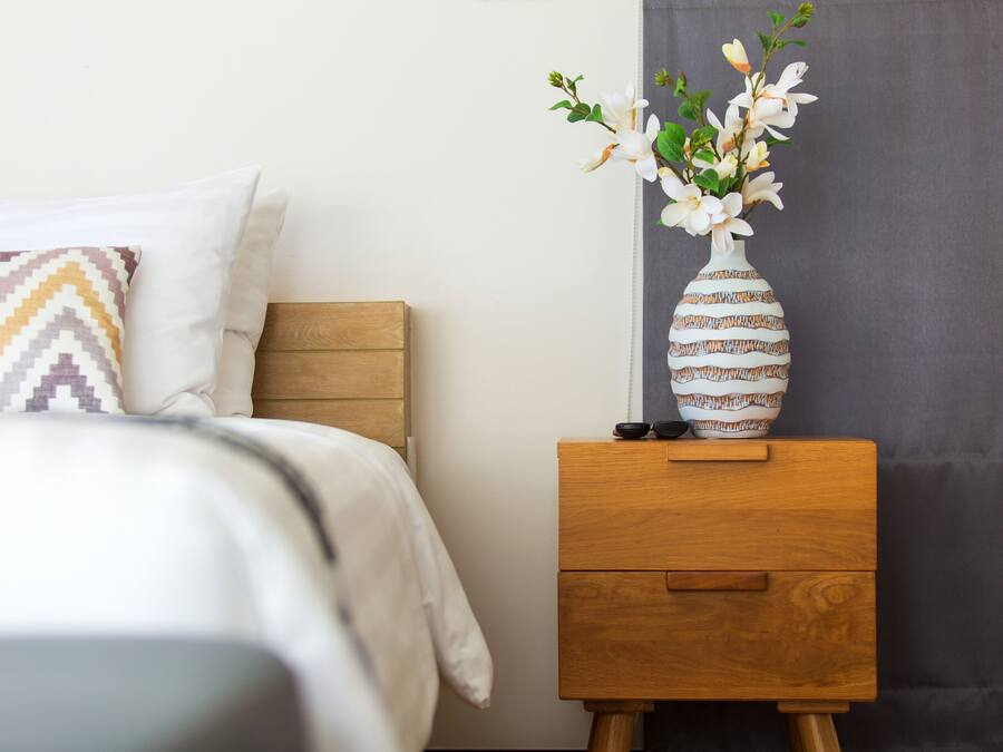 Una cama bien hecha con un cojín estampado, una mesita de noche con un florero y una cortina oscura.