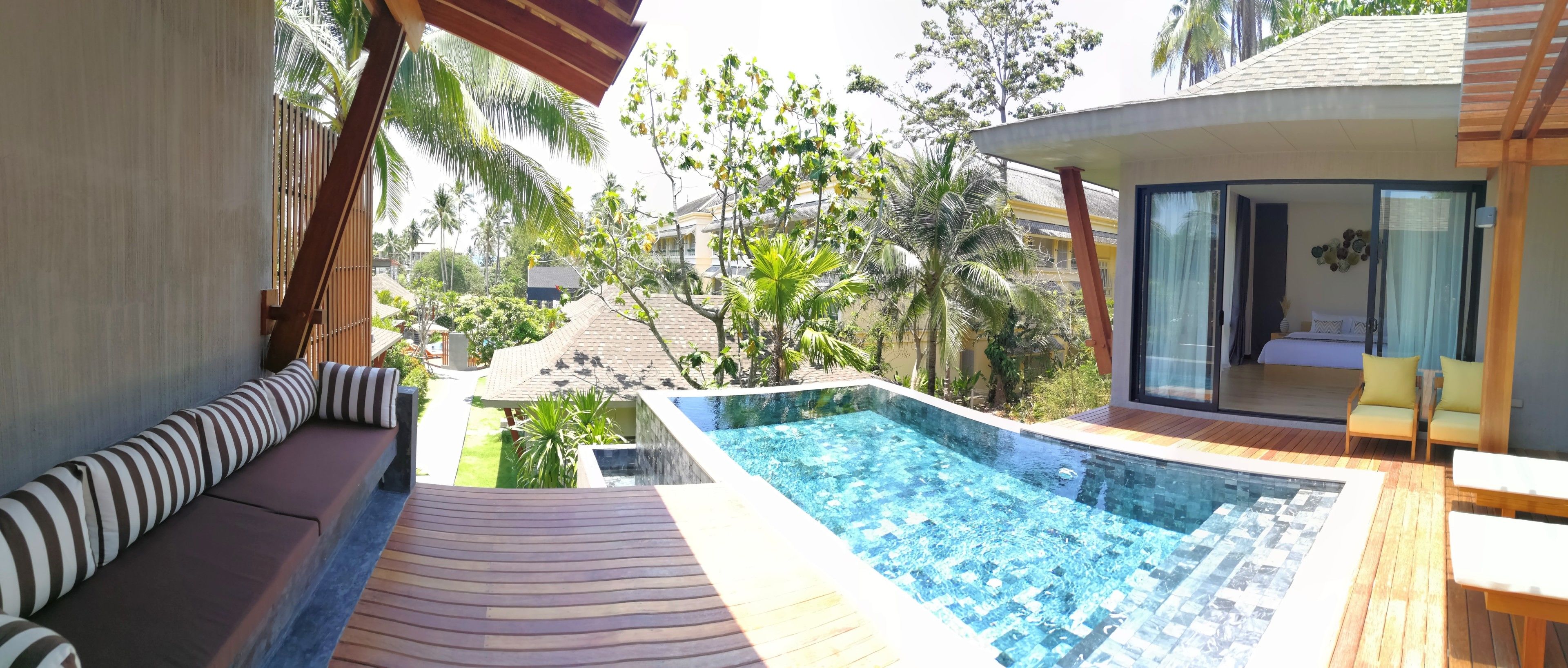 A pool area with a wooden deck, a seating area with striped cushions, and a view of surrounding buildings and greenery.