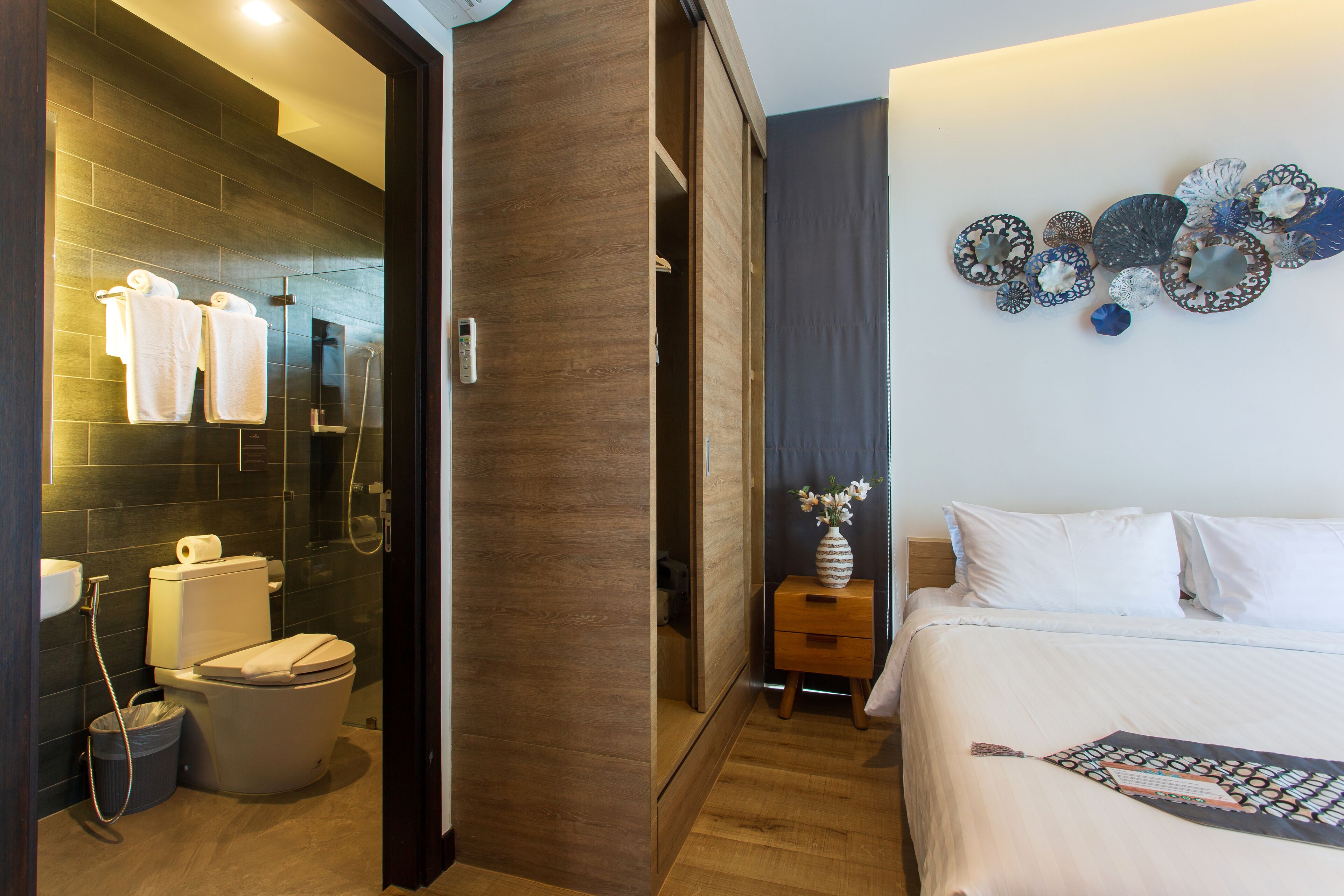 A hotel room with a wooden floor, a bed with white linens, a bedside table with a vase of flowers, a wall-mounted decorative plate, and a shower area with a glass door.