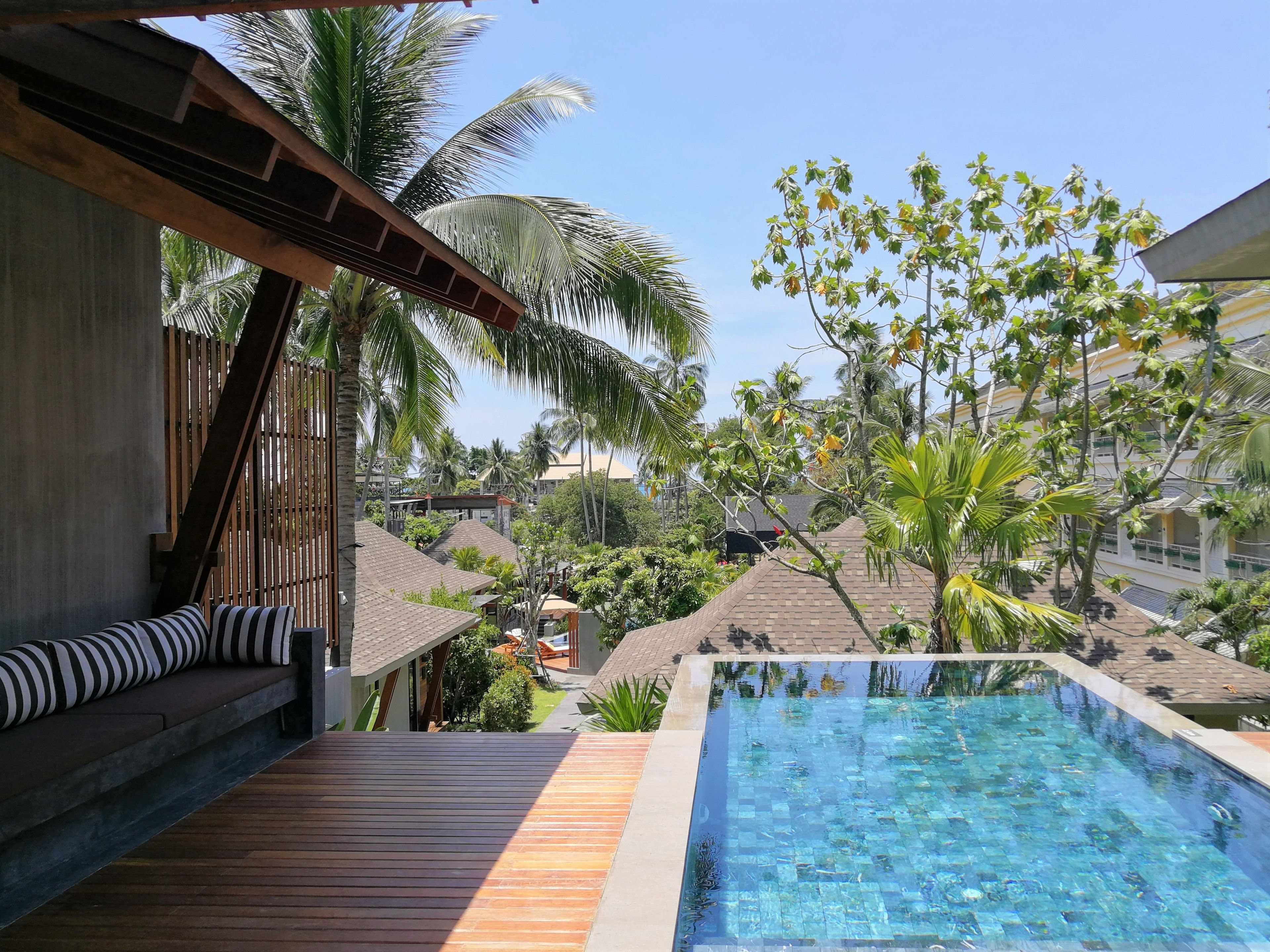 A pool area with wooden deck and lounge seating, surrounded by palm trees and a view of other buildings.