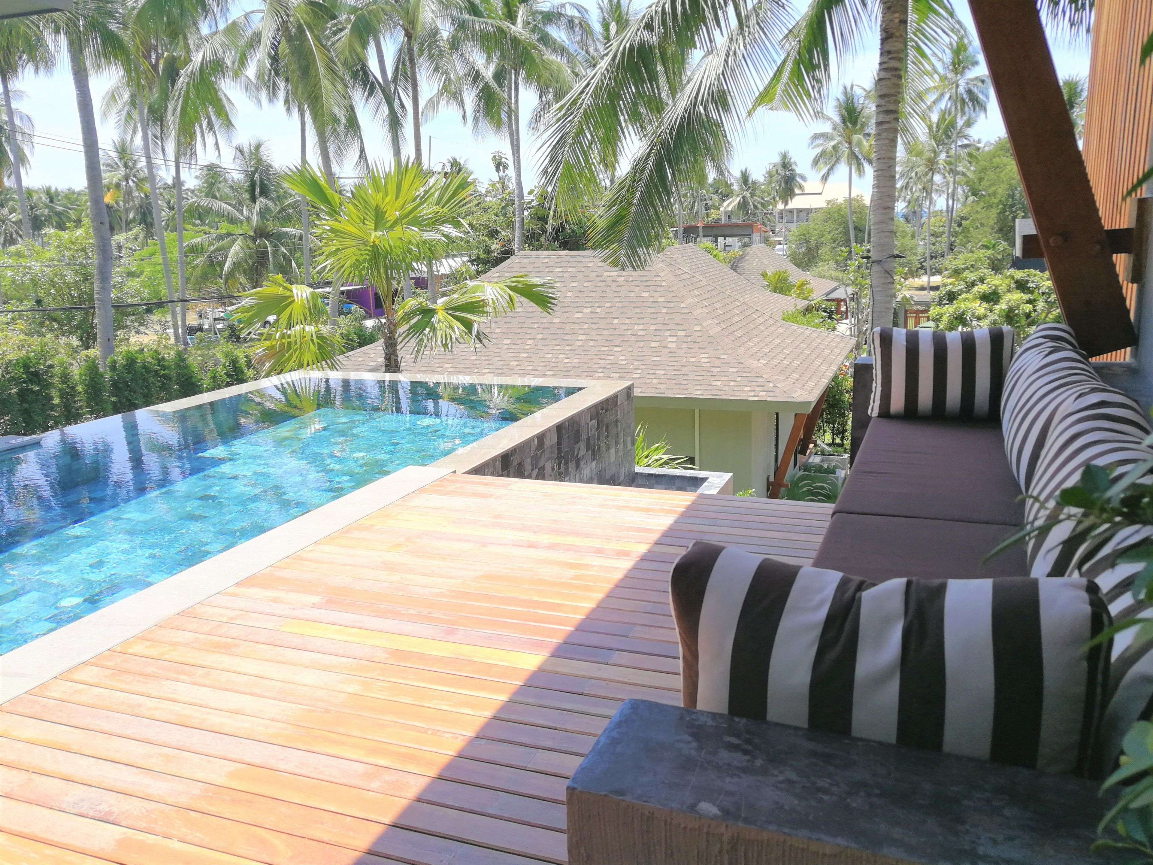 Una terraza de madera con piscina, zona de descanso con cojines a rayas y vistas a palmeras y edificios.