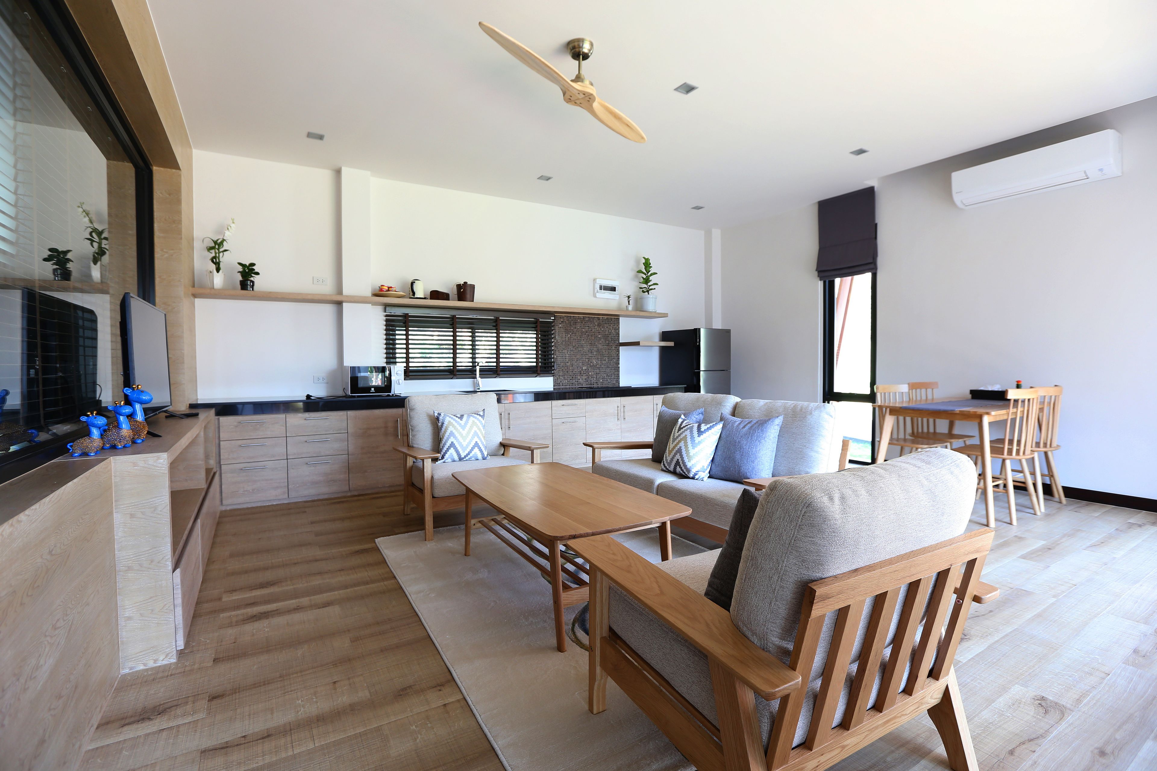 A modern living room with a wooden sofa, a coffee table, and a dining area with a wooden table and chairs. There is a kitchen area with a refrigerator and a window with blinds.
