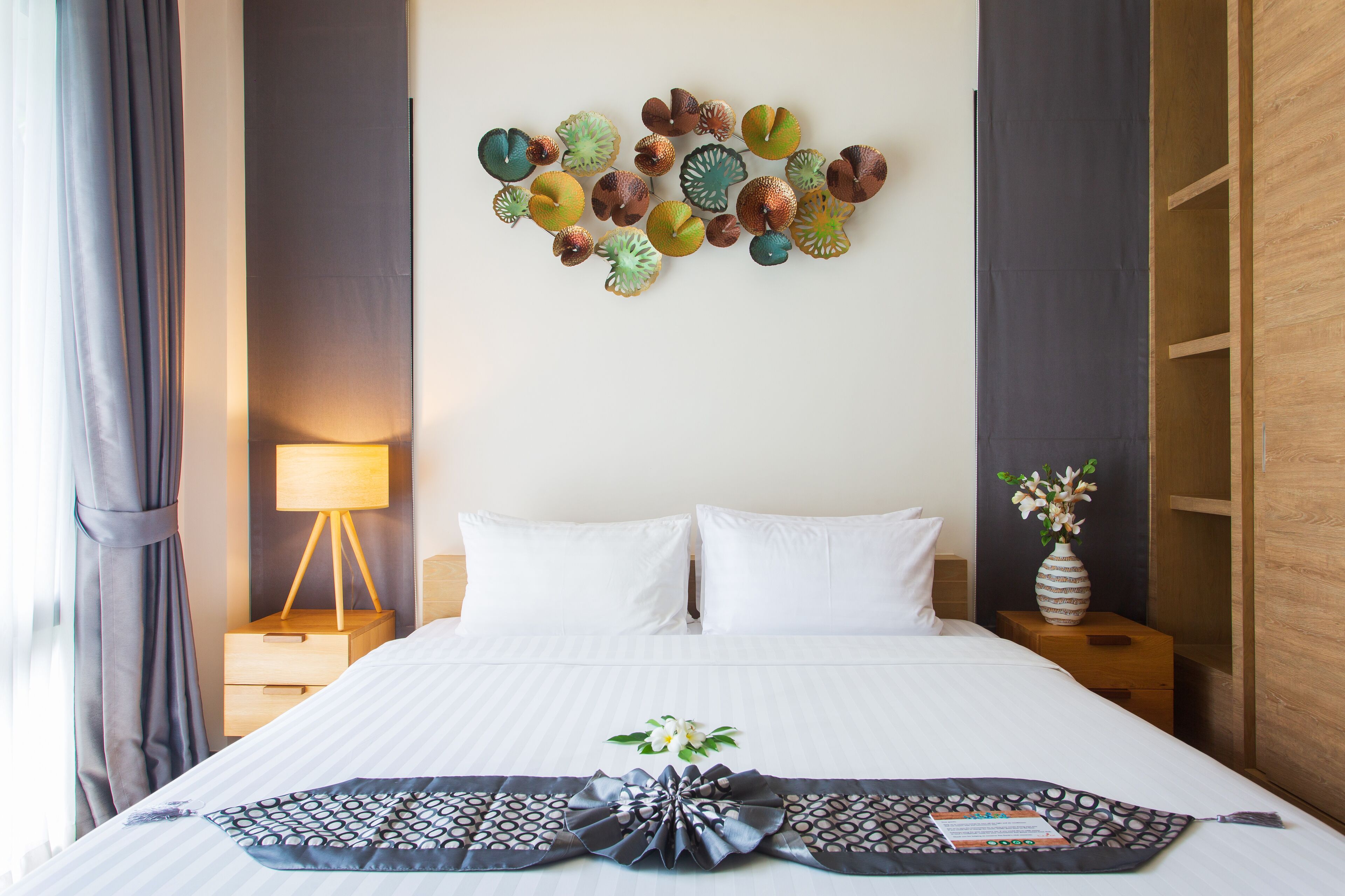 A neatly made bed with white linens, a decorative wall piece above it, a bedside table with a lamp, and a vase with flowers on the nightstand.