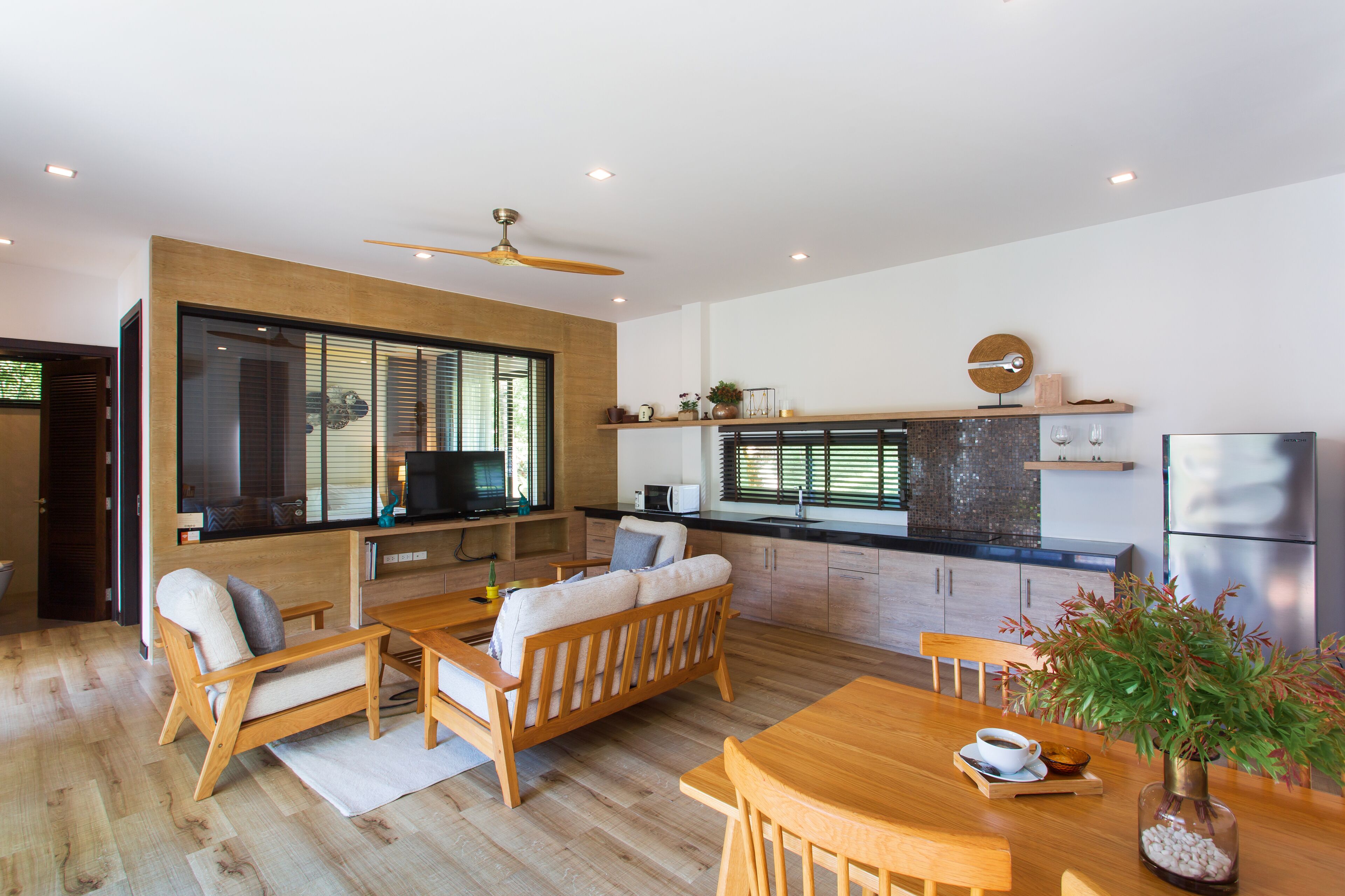 A modern kitchen with wooden cabinets, a dining area with a wooden table and chairs, and a large window with blinds.