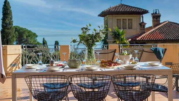 Un espace extérieur aménagé pour les repas, avec une table dressée, entourée de chaises, et une vue sur un bâtiment doté d’un toit en tuiles.