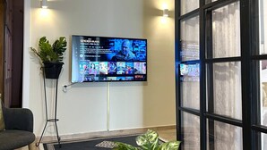 Un salon avec un téléviseur fixé au mur diffusant différentes émissions, une plante en pot, un lampadaire et une grande fenêtre avec des rideaux.