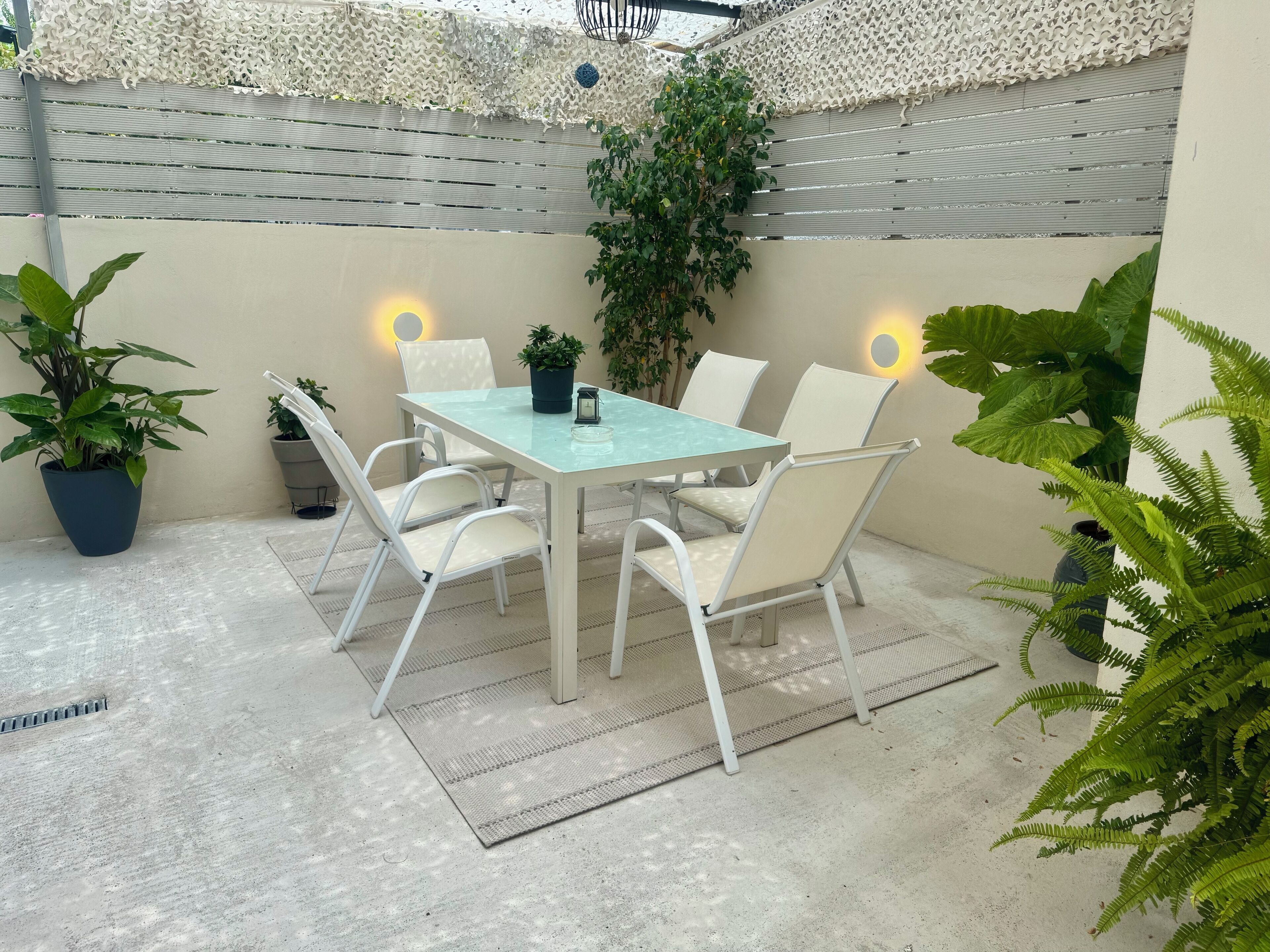 Un espacio de patio con mesa de vidrio y sillas blancas, rodeado de plantas en macetas y un enrejado blanco decorativo.