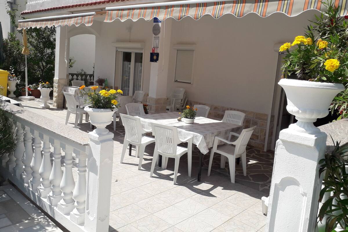 Um pátio externo com cadeiras e mesas de plástico branco, rodeado por balaústres brancos e vasos com flores amarelas.