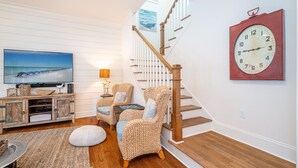 Un escalier avec des marches en bois et une rampe blanche, un téléviseur à écran plat posé sur un meuble en bois, des chaises en osier, un pouf rond et une horloge murale.