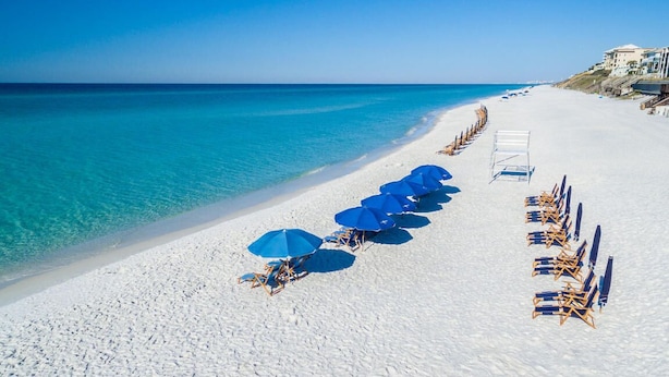 Une plage de sable blanc, avec une eau d’un bleu limpide et bordée de rangées de parasols bleus et de chaises de plage.