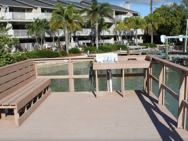 Een houten terras met een bankje, een kleine tafel met een gootsteen, en uitzicht op een jachthaven met boten.