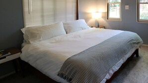 A neatly made bed with white linens and a striped throw blanket, a bedside lamp, and a nightstand with a book and a cup.