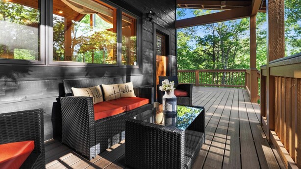 A covered outdoor seating area with wicker furniture and a glass table, surrounded by wooden railings and large windows. - Luxury Cabin | Hot Tub & Game Room Near Highlands (Franklin)