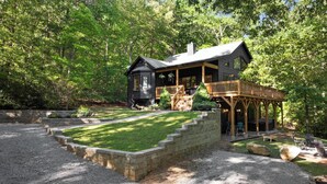A cabin with a deck, surrounded by a well-maintained lawn and a stone retaining wall. - Luxury Cabin | Hot Tub & Game Room Near Highlands (Franklin)
