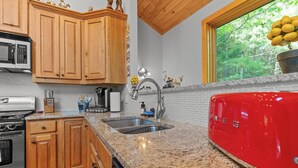 A kitchen with wooden cabinets, granite countertops, a red refrigerator, and a window with a view of trees. - Luxury Cabin | Hot Tub & Game Room Near Highlands (Franklin)