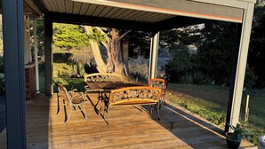 An outdoor wooden deck with a table and chairs, surrounded by a covered area with a view of a garden and a large tree. - River Front Estate (Huonville)