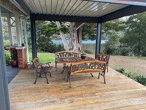 A covered outdoor patio with a wooden table and chairs, surrounded by greenery and a view of a lake. - River Front Estate (Huonville)