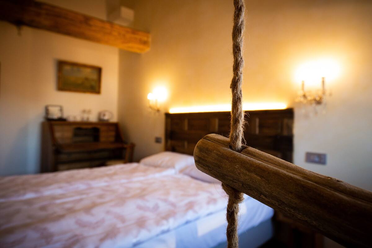 A bedroom with a wooden headboard, a bed with a patterned bedspread, a nightstand with a lamp, and a wall-mounted shelf with decorative items.