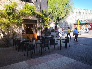 Un café al aire libre con mesas y sillas, un muro de piedra y una calle al fondo.