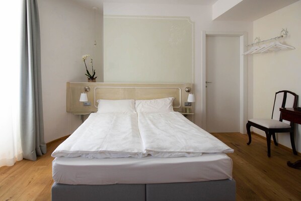 Una cama bien hecha con sábanas blancas, una mesita de noche con una lámpara y una silla a la derecha.