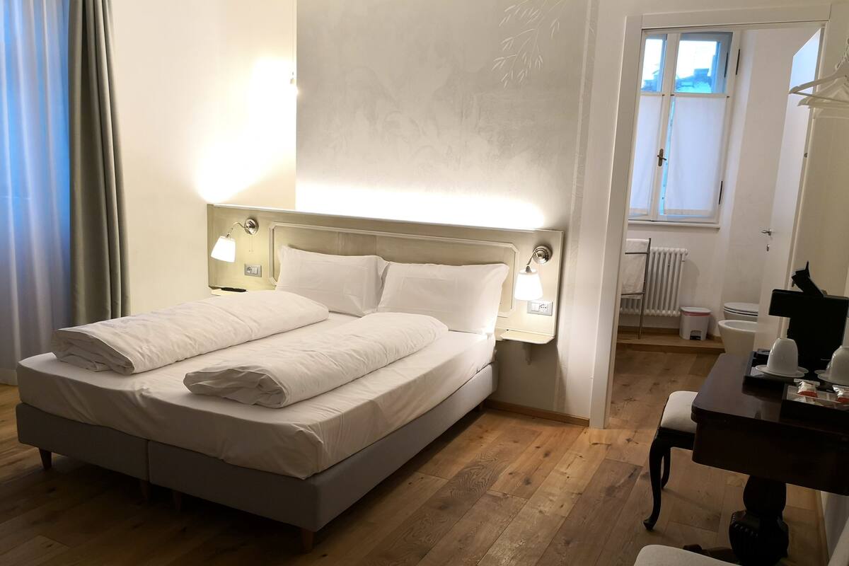 A neatly made bed with white linens, a bedside table with a lamp, and a wooden floor.