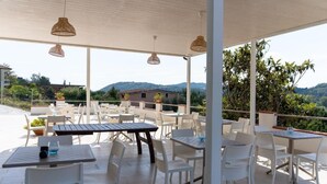 Un patio al aire libre con mesas y sillas blancas, lámparas colgantes de mimbre y vistas del paisaje circundante.