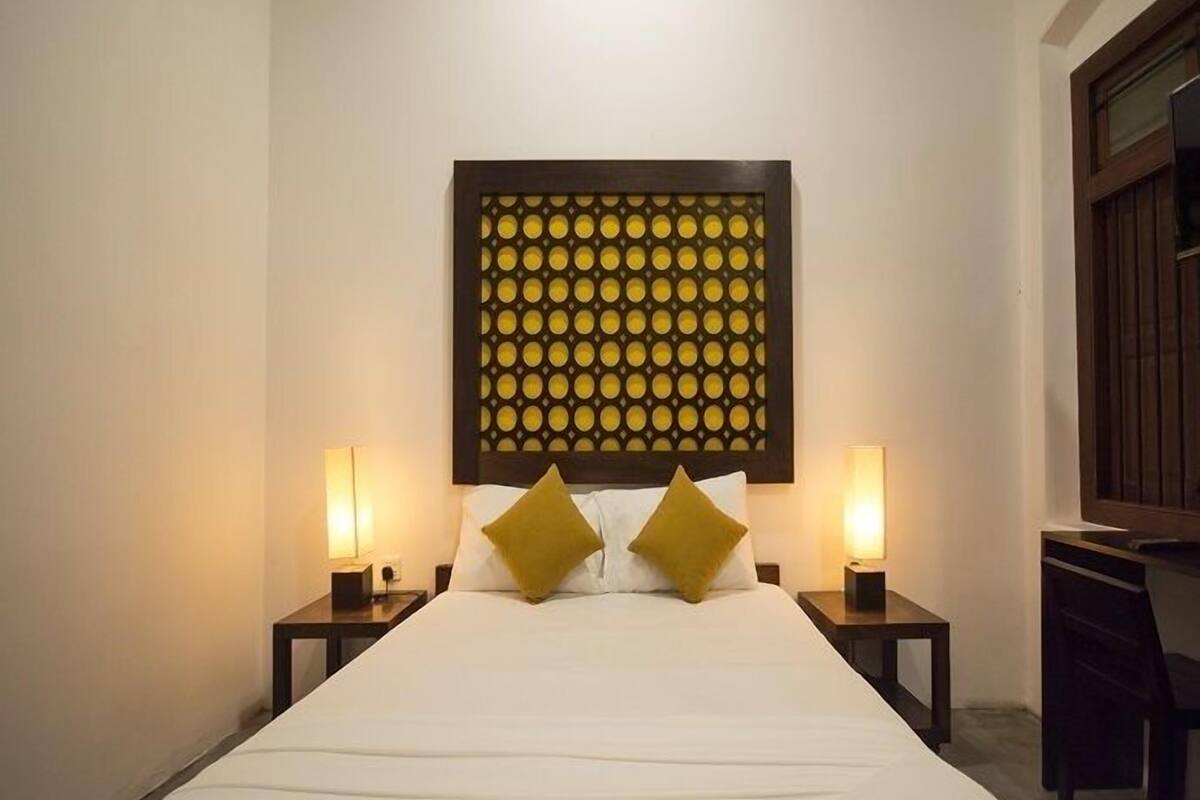 Una habitación de hotel con una cama grande, dos mesitas de noche, una obra de arte colgada en la pared y una ventana con persianas.