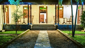 A pathway leading to a building with a wooden door, flanked by rocking chairs and a small tree. - Thambu Illam (Jaffna)
