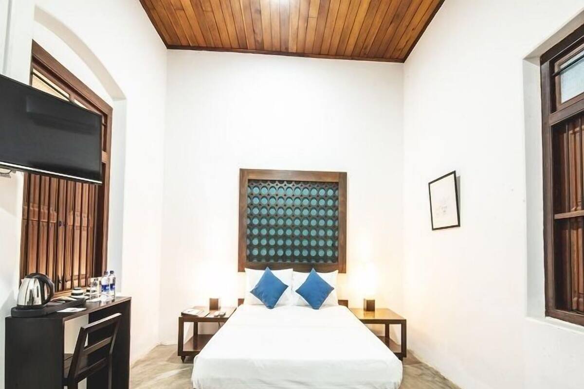 Un dormitorio con cabecera de madera, cama con ropa de cama blanca, dos mesitas de noche, un escritorio con silla y una ventana con contraventanas de madera.