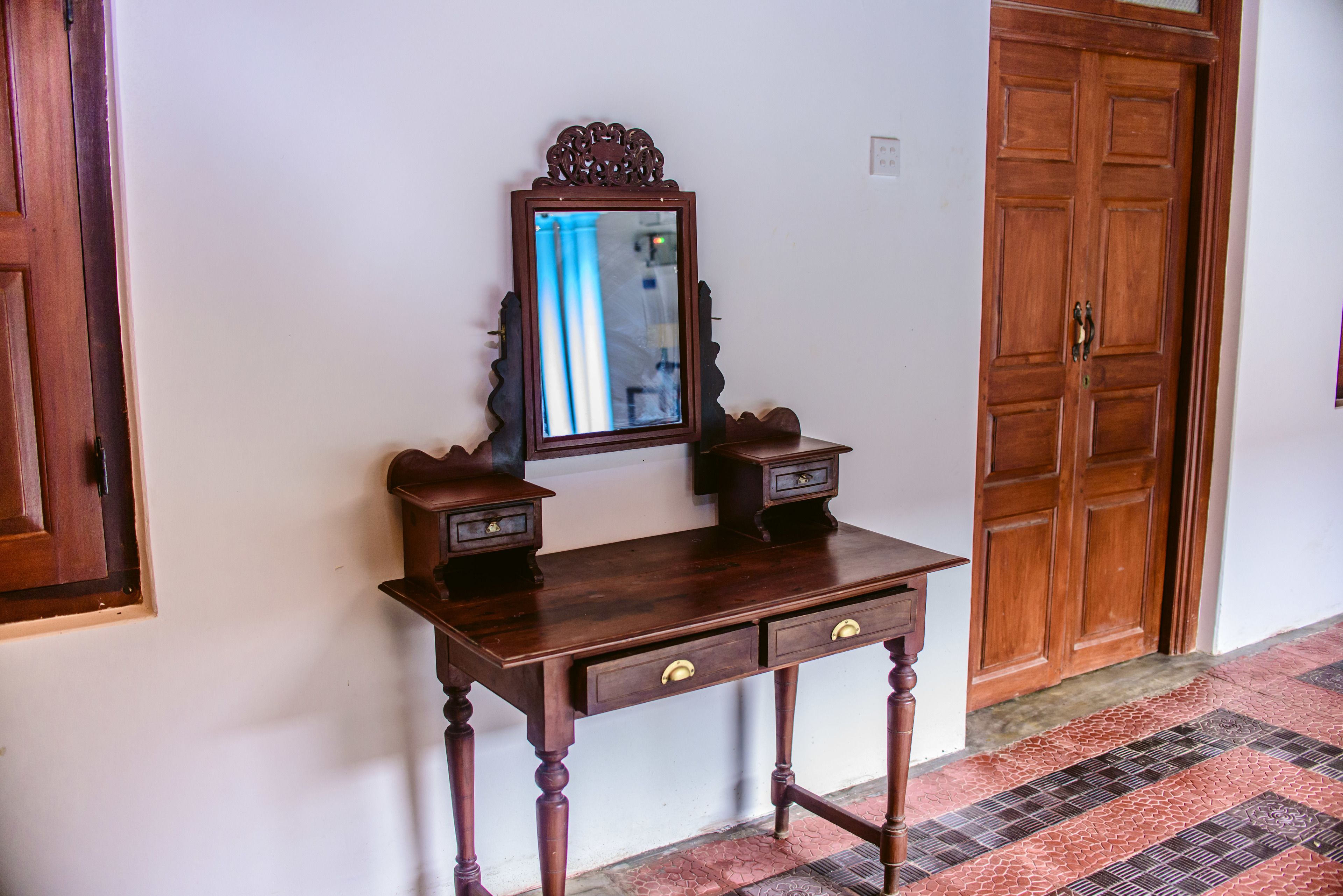 A wooden desk with two drawers and a mirror, positioned against a white wall with a wooden door in the background.
