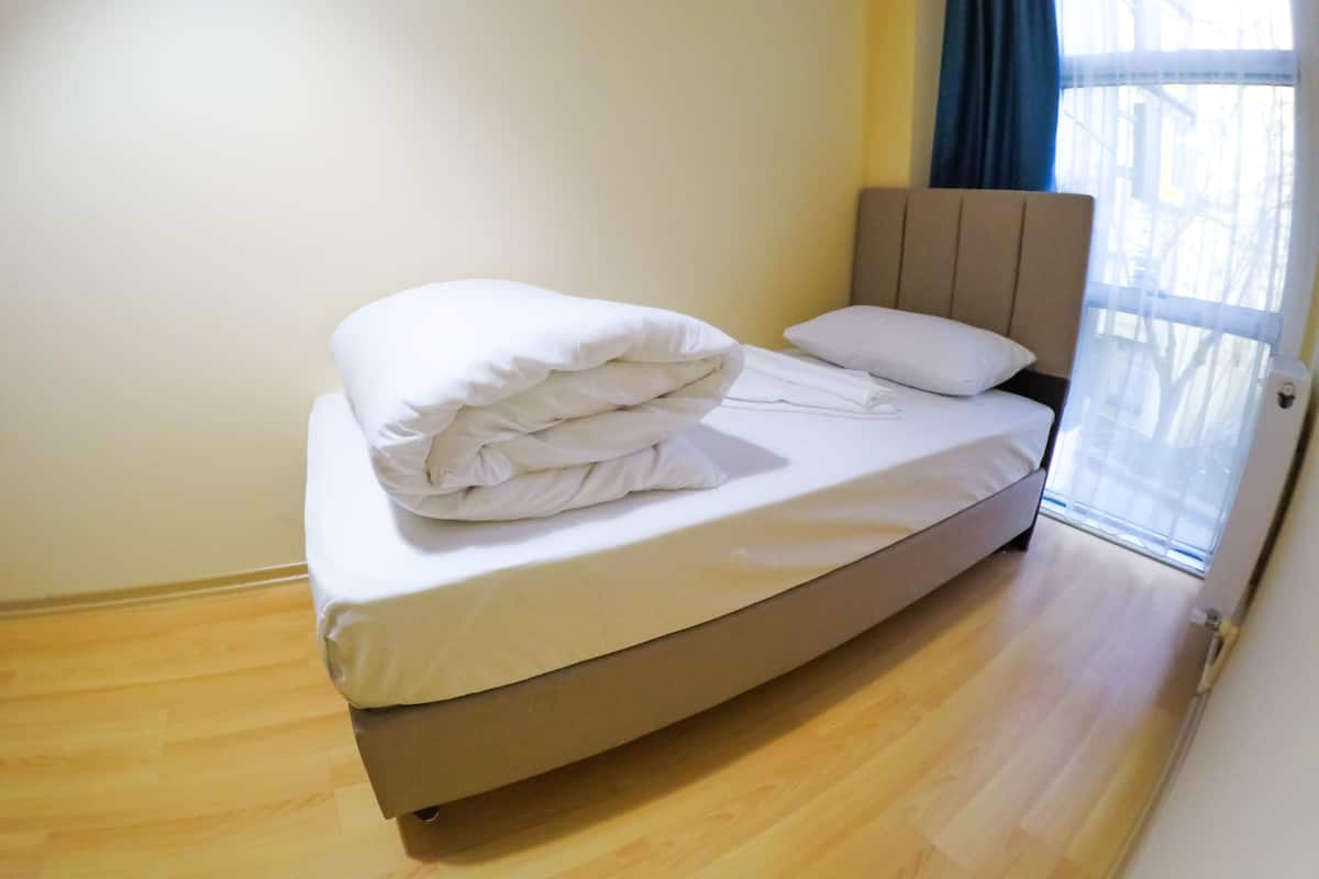 A single bed with a beige headboard and white bedding in a room with wooden flooring and a window with blue curtains.