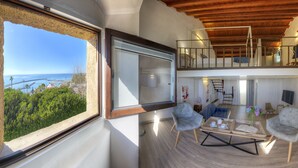 Amplia sala de estar con un ventanal que ofrece vistas al mar y al follaje, techo de madera y balcón con televisor.