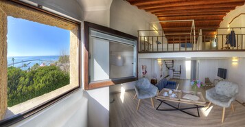 Un'ampia zona giorno con una grande finestra che offre vista sul mare e sul verde, un soffitto in legno e un balcone con televisore.