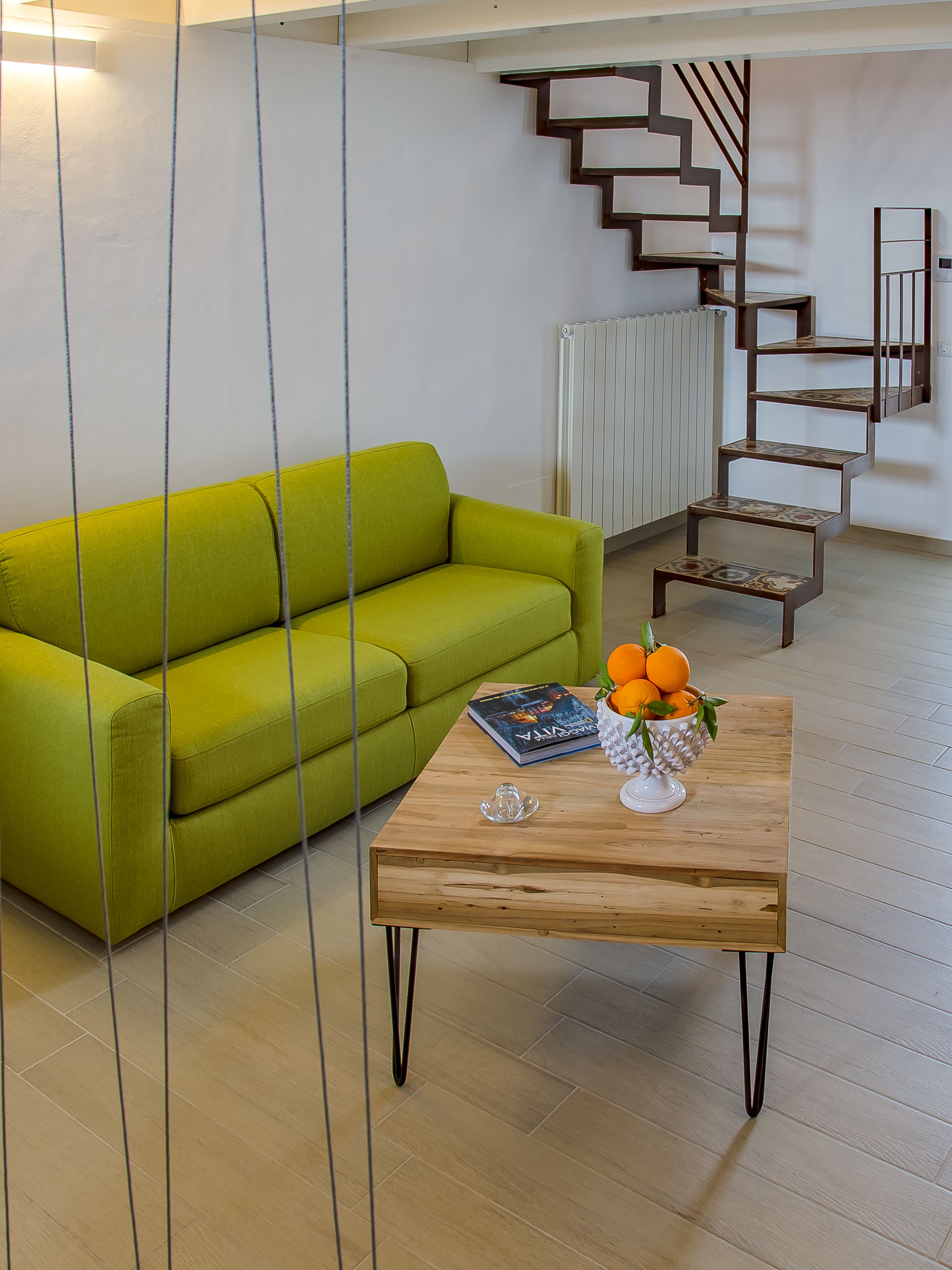 A modern living room with a green sofa, a wooden coffee table with a book and fruit bowl, and a staircase with a railing.