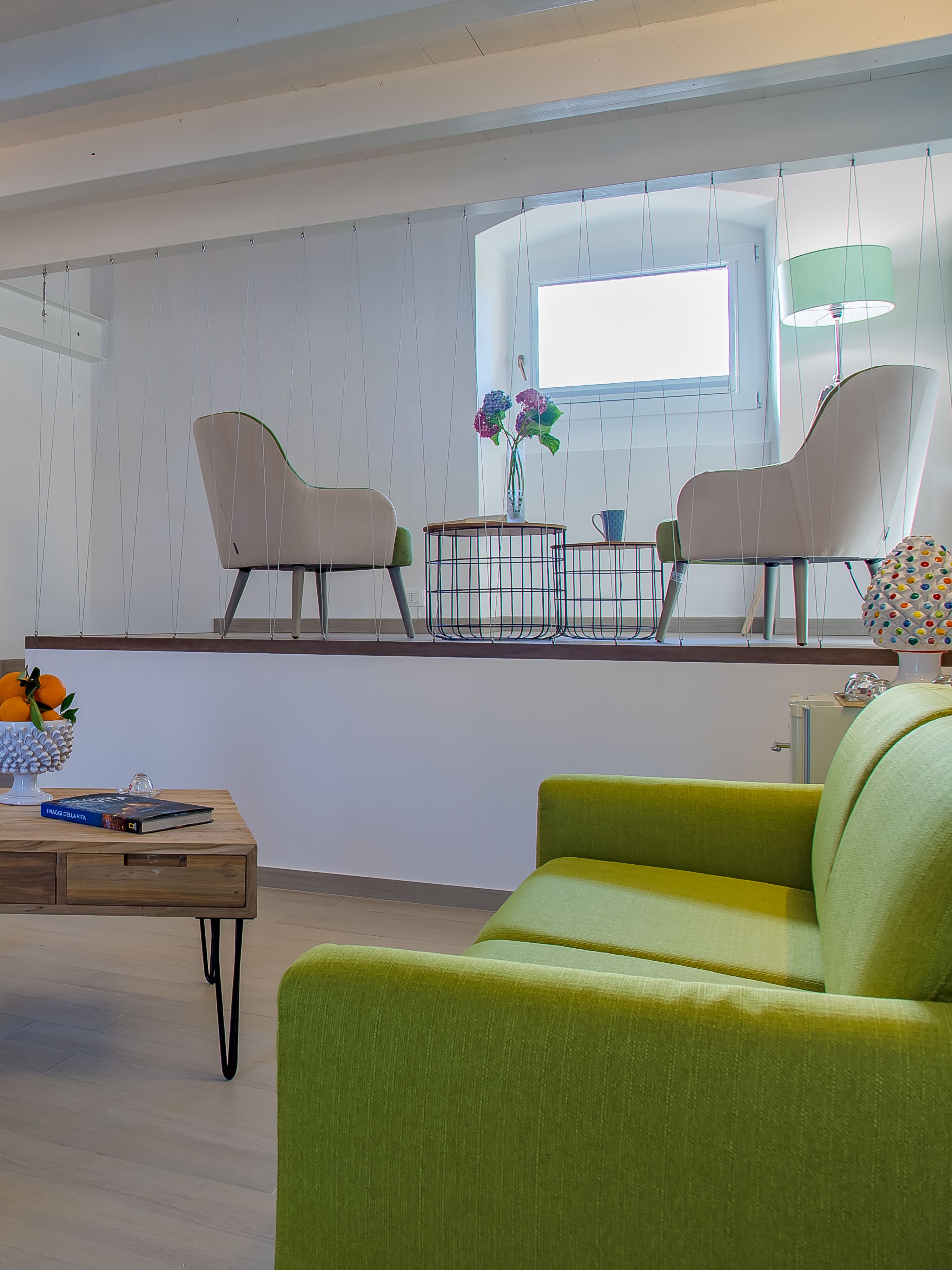 Un salón moderno con un sofá verde, dos sillones, una mesa de centro con libros y un jarrón de flores, y una ventana con cortinas.
