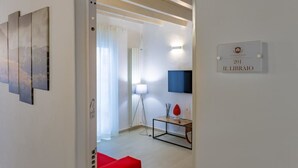 Una sala moderna con un sofá rojo, un televisor montado en la pared y una mesita con objetos decorativos. Un cartel en la pared indica el número y el nombre de la habitación.
