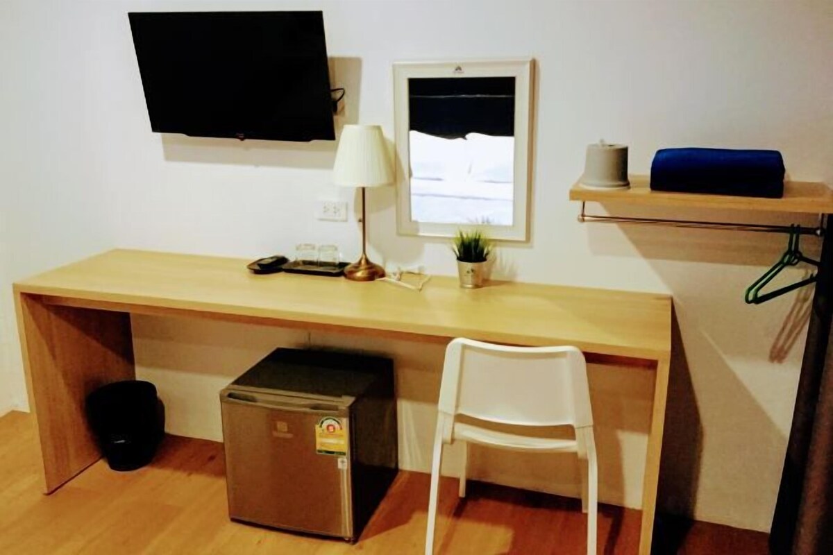 A compact room with a wooden desk, a white chair, a small fridge, a TV mounted on the wall, a lamp, a framed picture, and a shelf with a roll of paper towels.