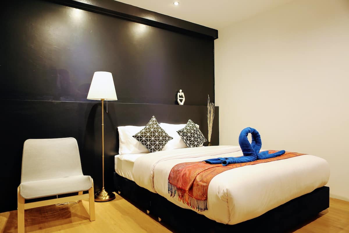 A neatly made bed with a white comforter, a blue towel, and patterned pillows. A white chair is positioned next to the bed, and a floor lamp is to the left.