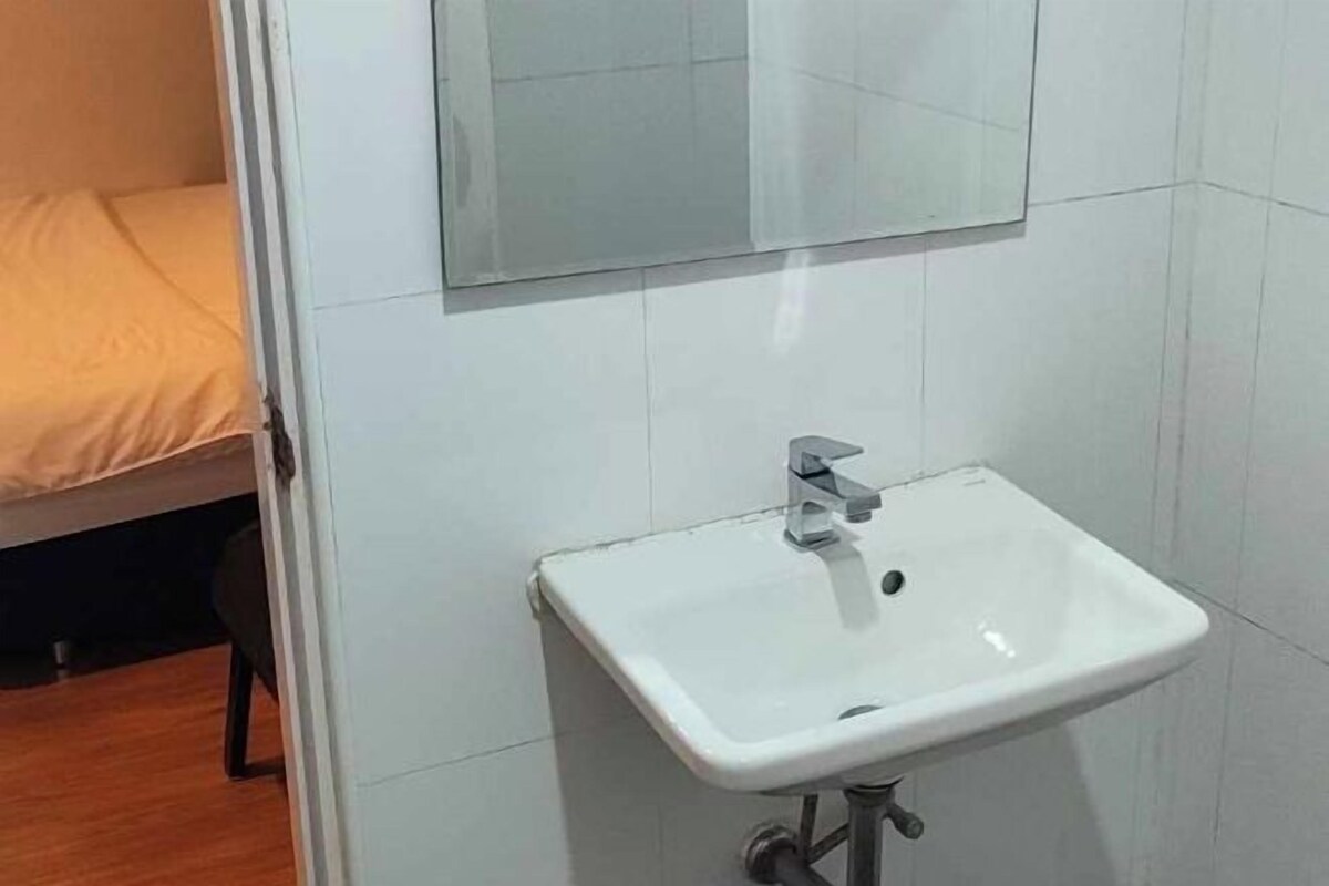 A bathroom with a white sink, a mirror above it, and a bed with a beige cover in the background.